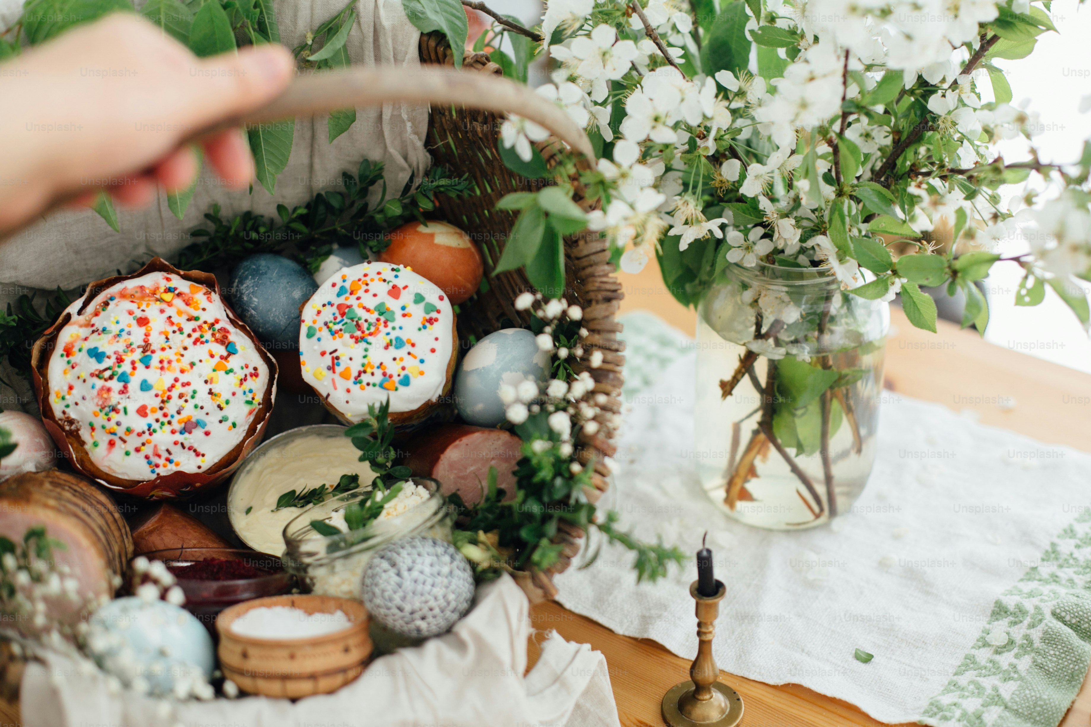 Traditioneller Osterkorb. Ostern moderne Eier, Osterbrot, Schinken, Rüben, Butter, Würstchen in rustikalem Korb dekoriert mit grünen Buchsbaumzweigen und Blumen auf Holztisch mit Kerze