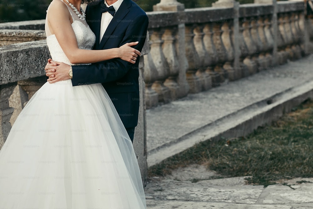 Elegante Braut und Bräutigam umarmen sich sanft. Hochzeitspaar umarmt sich am Abend im alten Schloss. Platz für Text. Mann im klassischen Anzug mit Fliege und Frau im weißen Kleid mit Perlen