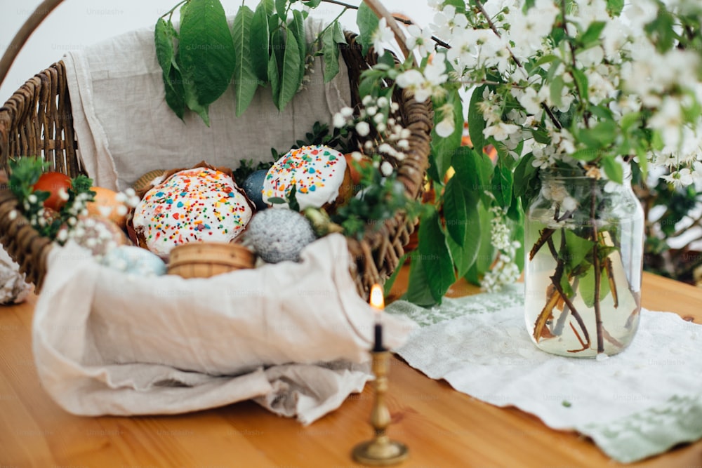 伝統的なイースターバスケット。イースターモダンエッグ、イースターパン、ハム、ビート、バター、キャンドル付きの木製のテーブルの上に緑のツゲの枝と花で飾られた素朴なバスケットに入ったソーセージ