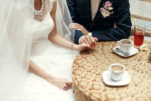 stylish wedding couple holding hands, sitting at wooden vintage table with warm coffee in cups in cafe. happy  bride and groom in dress and suit  hugging