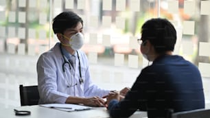 Cropped image of doctor talking/discussing with young patient man while sitting together at the doctor working desk with examination room as background.