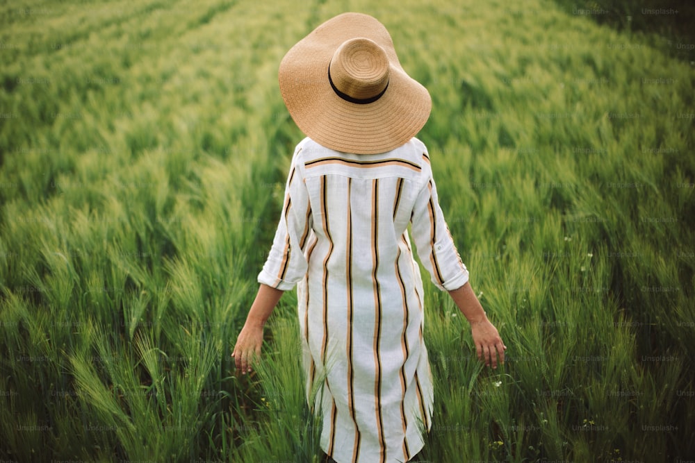 Woman in rustic dress and hat walking in green field of barley. Atmospheric authentic moment. Stylish girl enjoying peaceful evening in countryside. Copy space. Rural slow life