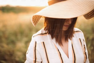 Retrato da mulher bonita no chapéu apreciando a luz dourada do pôr do sol no prado do verão. Menina rústica elegante no vestido de linho relaxando à noite no campo. Vida lenta rural. Momento atmosférico