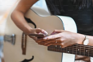 Cropped image of sexy brunette woman using smartphone while playing an acoustic guitar and sitting at the modern chair with modern/orderly music studio as background.