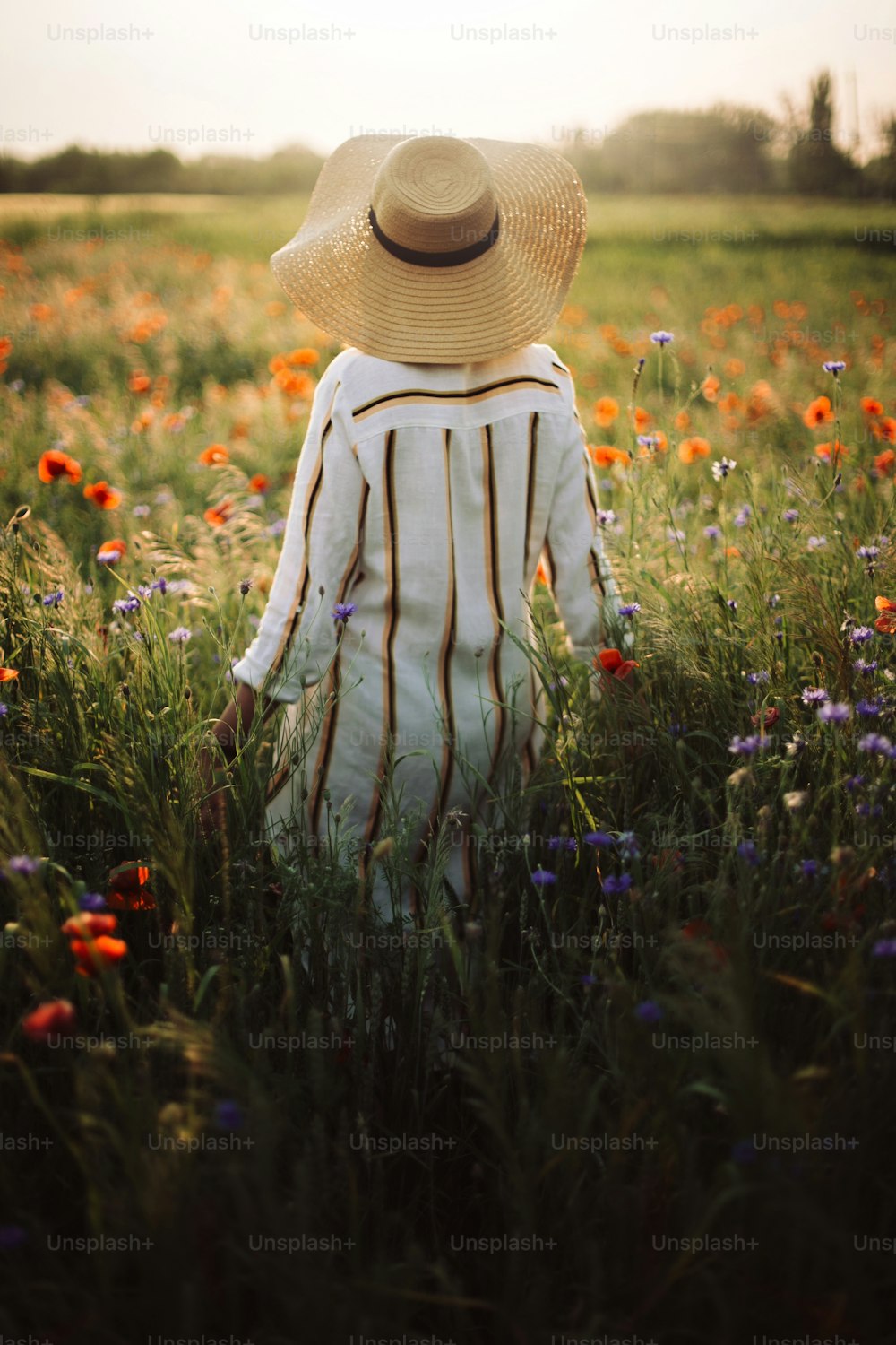 Junge Frau in rustikalem Leinenkleid spaziert zwischen Mohn und Kornblumen in sommerlicher Wiesenlandschaft im Sonnenuntergangslicht. Ländliches langsames Leben. Das einfache Leben genießen. Mädchen mit Hut in Wildblumen