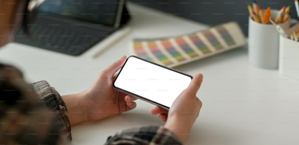 Foto recortada de una diseñadora sosteniendo un teléfono inteligente de maqueta horizontal mientras trabaja con una tableta y suministros de diseñador en una mesa blanca