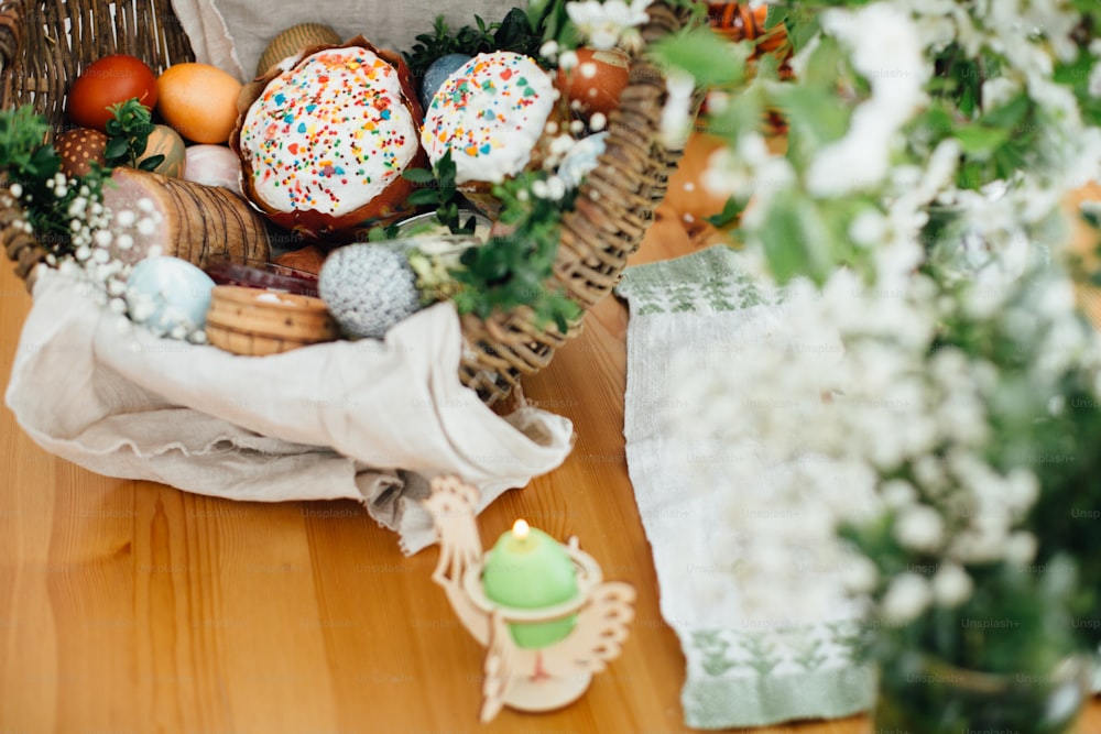 Cesta tradicional de Pascua para las bendiciones en la iglesia. Huevos modernos de Pascua, pastel, jamón, remolacha, mantequilla en cesta rústica decorada con ramas de buxus verde y flores en mesa de madera con vela