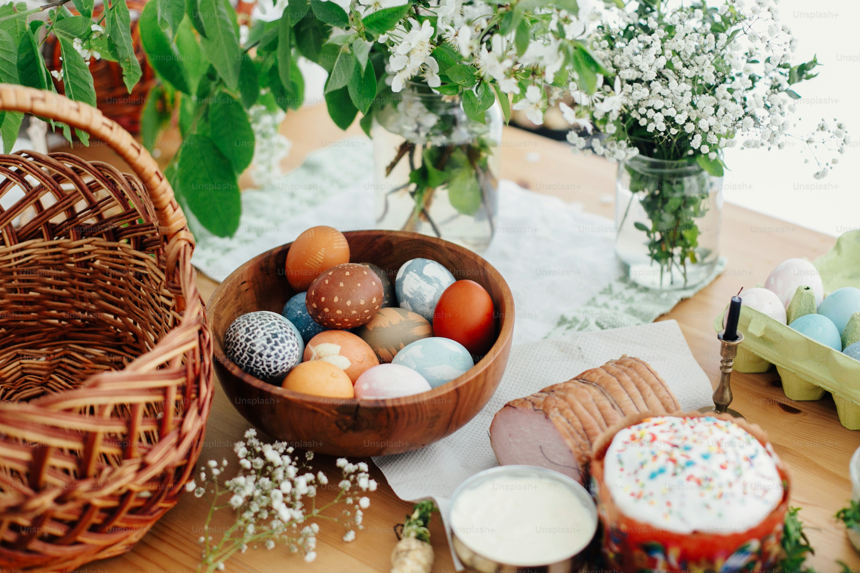 Ostereier natur gefärbt, Osterbrotkuchen, Schinken, Butter, grüne Zweige und Blumen auf rustikalem Holztisch mit Weidenkorb und Kerze. Traditionelles Osteressen zum Segen in der Kirche