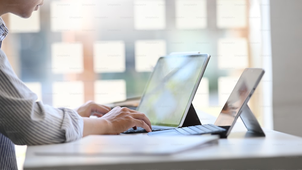 Imagen recortada de hombre de negocios usando/escribiendo en una tableta de computadora con una funda de teclado mientras está sentado en la mesa de trabajo moderna con un lugar de trabajo ordenado como fondo.