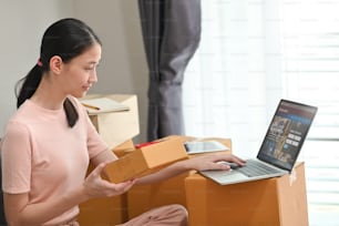 Foto einer jungen schönen Frau, die ihre Waren für die Lieferung an den Kunden mit Computer-Tablet und Laptop im gemütlichen Wohnzimmer verpackt. Start-up/Entrepreneur-Konzept.
