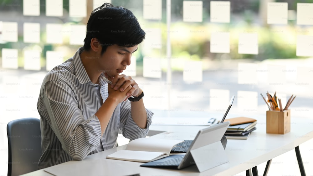 プロジェクト管理者として働くスマートな男性は、現代のオフィスを背景に、キーボードケース付きのコンピュータータブレットの前に座りながら、ノートブックに集中しています