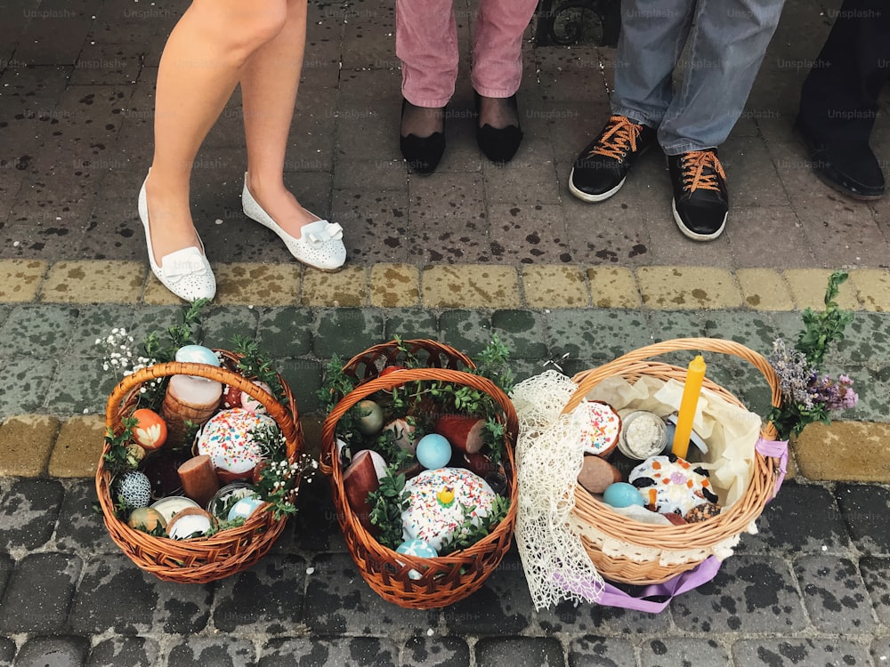 Easter baskets with stylish painted eggs, easter cake, ham,beets, butter, candle with boxwood branches for sanctify at church on background of standing people feet. Easter food for blessing