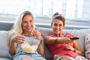 Portrait of funny and happy young women watching comedy in bed and laughing. Cheerful friends eating tasty popcorn and looking movie with gladness. Cozy and friendly atmosphere