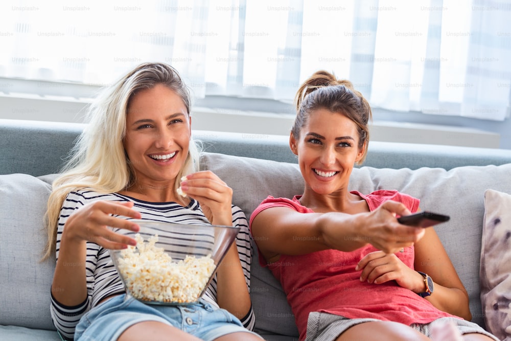 Portrait of funny and happy young women watching comedy in bed and laughing. Cheerful friends eating tasty popcorn and looking movie with gladness. Cozy and friendly atmosphere