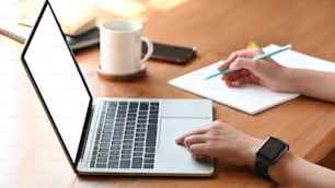 Cropped image of secretary woman's hands while typing on white blank screen computer laptop and writing on notebook at the modern wooden table with coffee cup, smartphone and notebook putting on it.