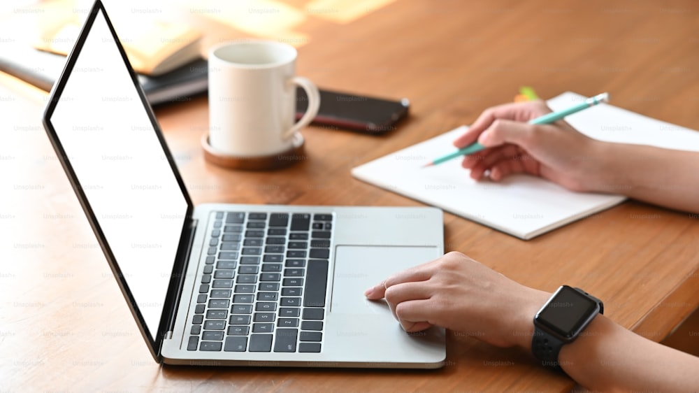 Cropped image of secretary woman's hands while typing on white blank screen computer laptop and writing on notebook at the modern wooden table with coffee cup, smartphone and notebook putting on it.