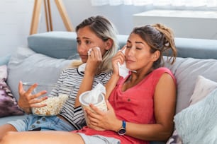 two best friends watch sad movie at home in bed crying touching. girls hold popcorn and tv remote control looking screen romantic film on television. ladies in pajama wiping tear using tissue