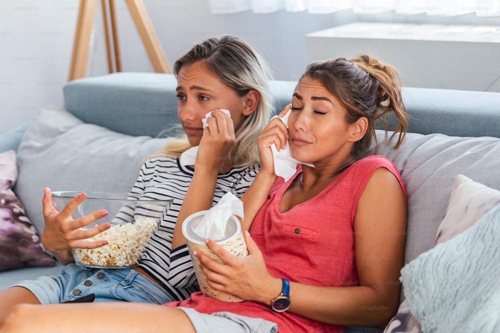 two best friends watch sad movie at home in bed crying touching. girls hold popcorn and tv remote control looking screen romantic film on television. ladies in pajama wiping tear using tissue