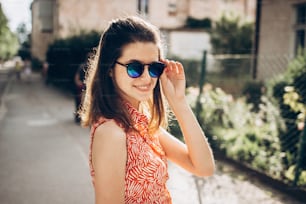 세련된 힙스터 여자는 선글라스를 끼고 웃으며 여름에 햇살이 내리쬐는 거리에서 햇살을 즐기고 있다. 어린 소녀가 포즈를 취하고 웃고 있다. 텍스트 공간. 즐거운 순간. 여름 여행