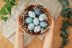 Hands holding wooden bowl with modern Easter eggs in spring flowers and eucalyptus on rustic table. Stylish pastel blue Easter eggs painted in natural dye from red cabbage. Happy Easter greetings