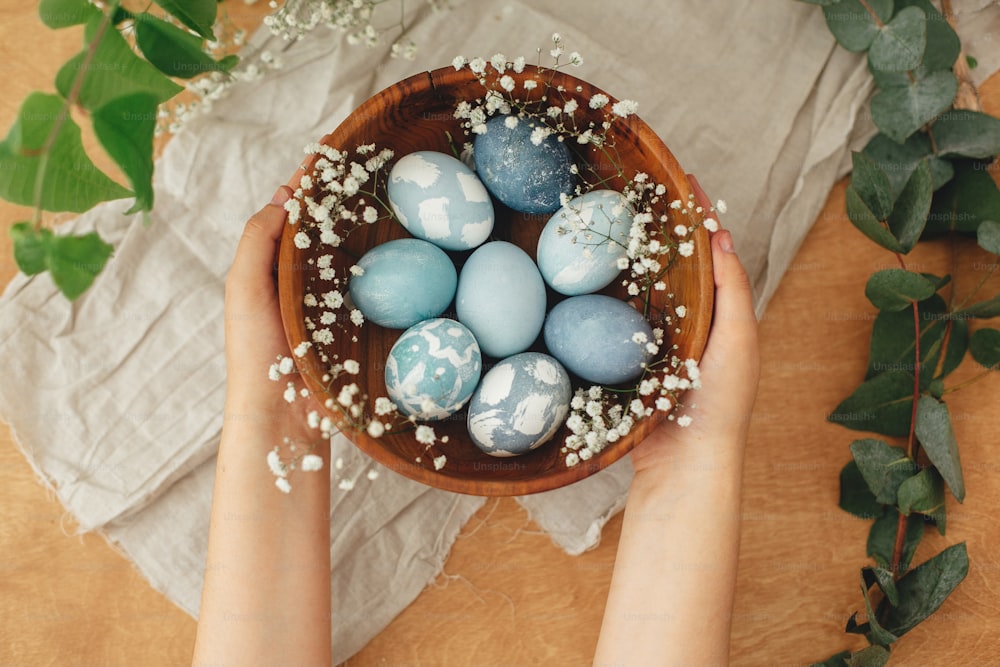 Hands holding wooden bowl with modern Easter eggs in spring flowers and eucalyptus on rustic table. Stylish pastel blue Easter eggs painted in natural dye from red cabbage. Happy Easter greetings