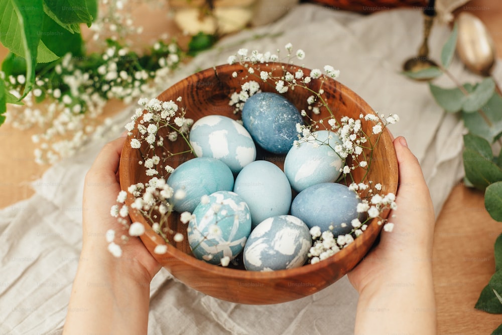 Frohe Ostern. Hände halten Holzschale mit modernen Ostereiern in Frühlingsblumen und Eukalyptus auf rustikalem Tisch. Stilvolle pastellblaue Ostereier, lackiert in natürlichem Farbstoff aus Rotkohl.