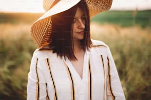Portrait of beautiful woman in hat enjoying sunset golden light in summer meadow. Stylish rustic girl in linen dress relaxing in evening in countryside. Rural slow living. Atmospheric moment