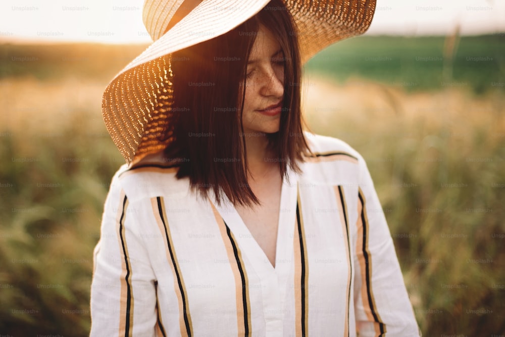 Portrait of beautiful woman in hat enjoying sunset golden light in summer meadow. Stylish rustic girl in linen dress relaxing in evening in countryside. Rural slow living. Atmospheric moment