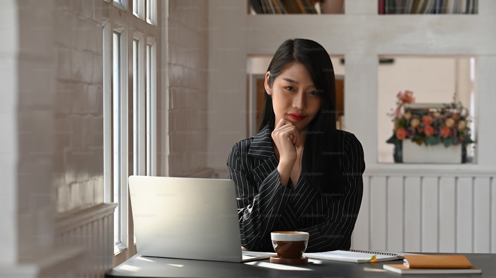 Beautiful woman in black striped suit working as business executive keep hand on chin while sitting at the modern working table with orderly workplace as background.