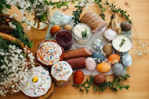 Traditional Easter Food. Easter modern eggs, easter bread, ham, beets, butter, cheese, sausages, green branches and flowers on rustic wooden table. Holiday preparation. Easter brunch