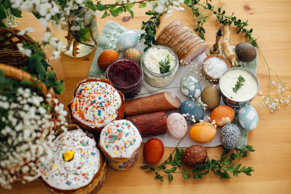 Traditional Easter Food. Easter modern eggs, easter bread, ham, beets, butter, cheese, sausages, green branches and flowers on rustic wooden table. Holiday preparation. Easter brunch