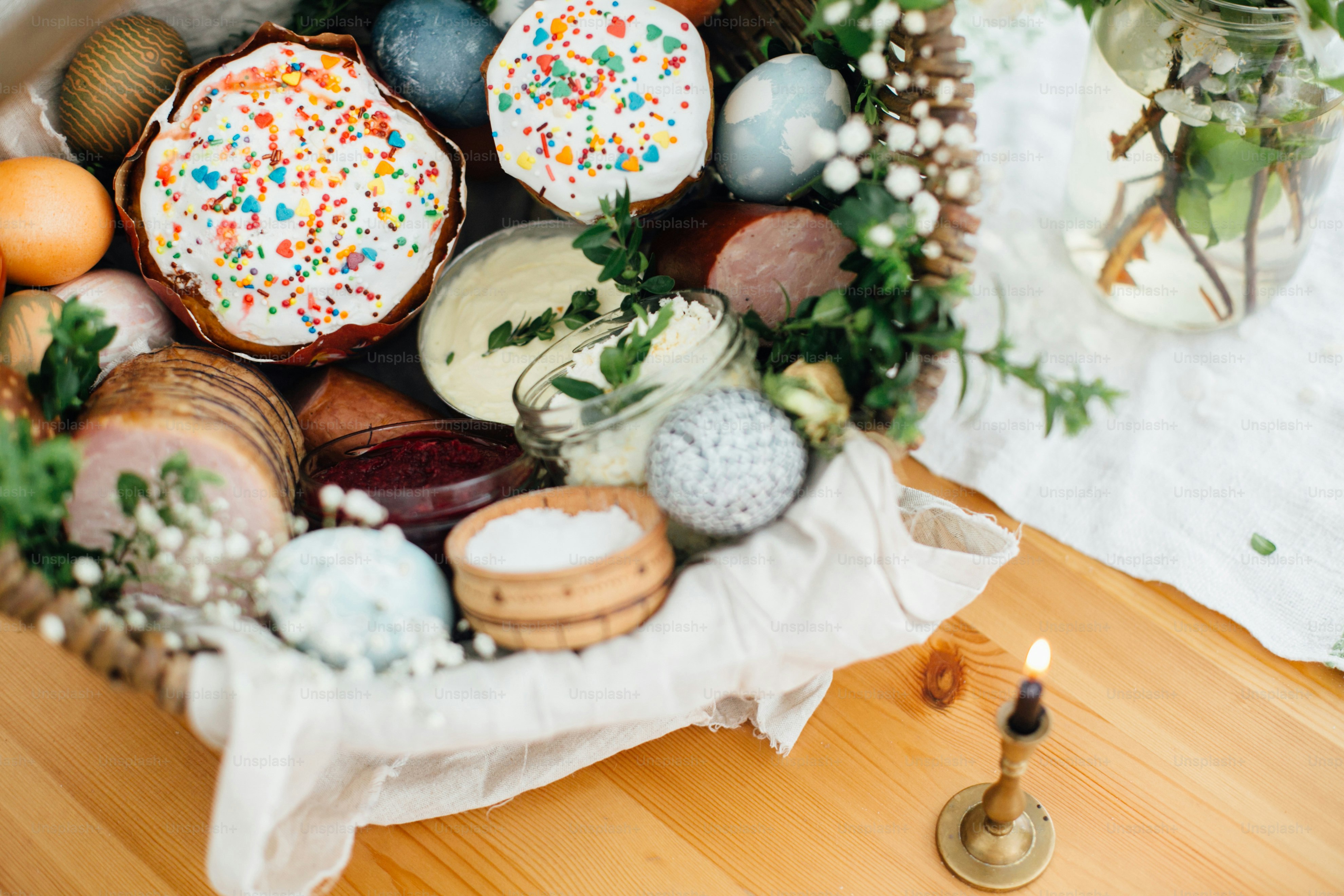 Ostermoderne Eier, Osterbrot, Schinken, Rüben, Butter, Würstchen im rustikalen Korb mit grünen Buchsbaumzweigen und Blumen auf Holztisch mit Kerze. Traditioneller Osterkorb.