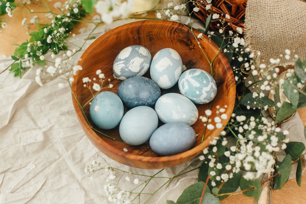 Modern Easter eggs in wooden bowl with spring flowers and eucalyptus on rustic table. Stylish pastel blue Easter eggs painted in natural dye from red cabbage. Happy Easter. Rural still life