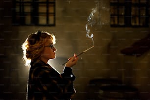Femme sexy élégante tenant une cigarette et fumant à l’extérieur, visage en gros plan, portrait de femme mystérieuse en manteau vintage, atmosphère noire française, concept de détective