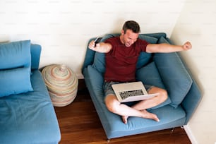 From above of joyful businessman in casual wear sitting on armchair with raised hand and opened mouth while working with laptop at home