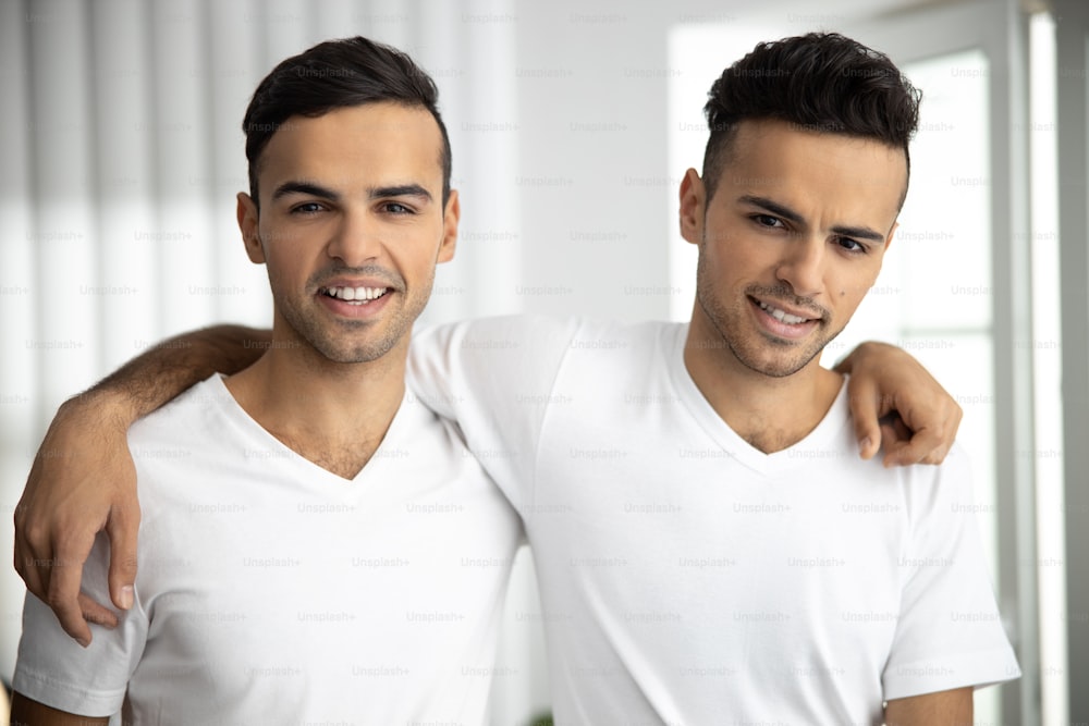 「自宅で抱きしめる白いTシャツを着た幸せでフレンドリーな男性の双子のウエストアップ」の写真素材(ストックフォト素材)