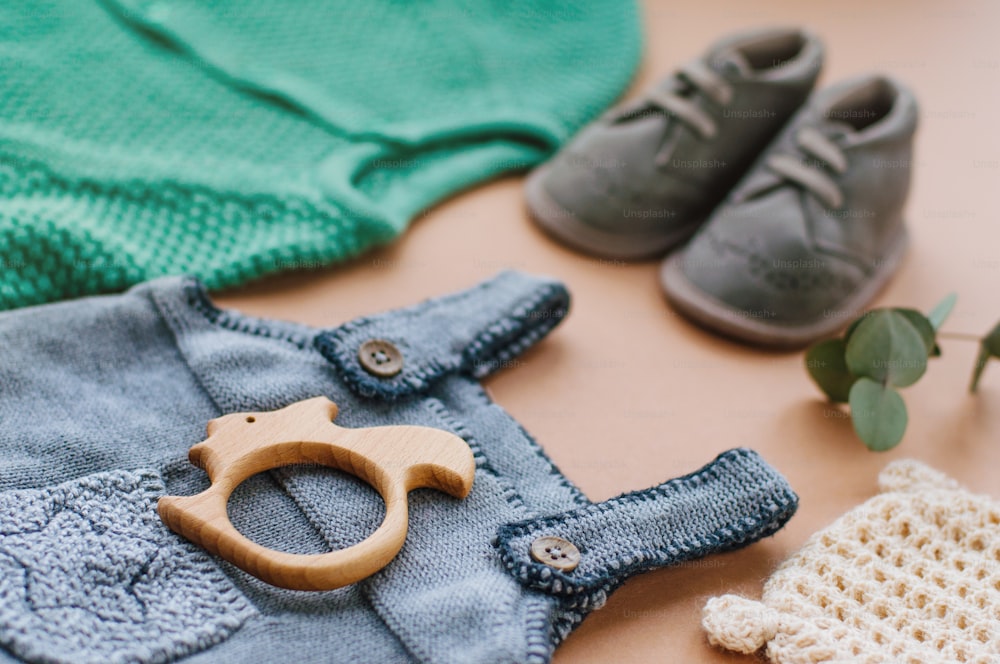 Baby clothes concept. Close up of baby knitted clothes and shoes on beige background near wooden teether squirrel.