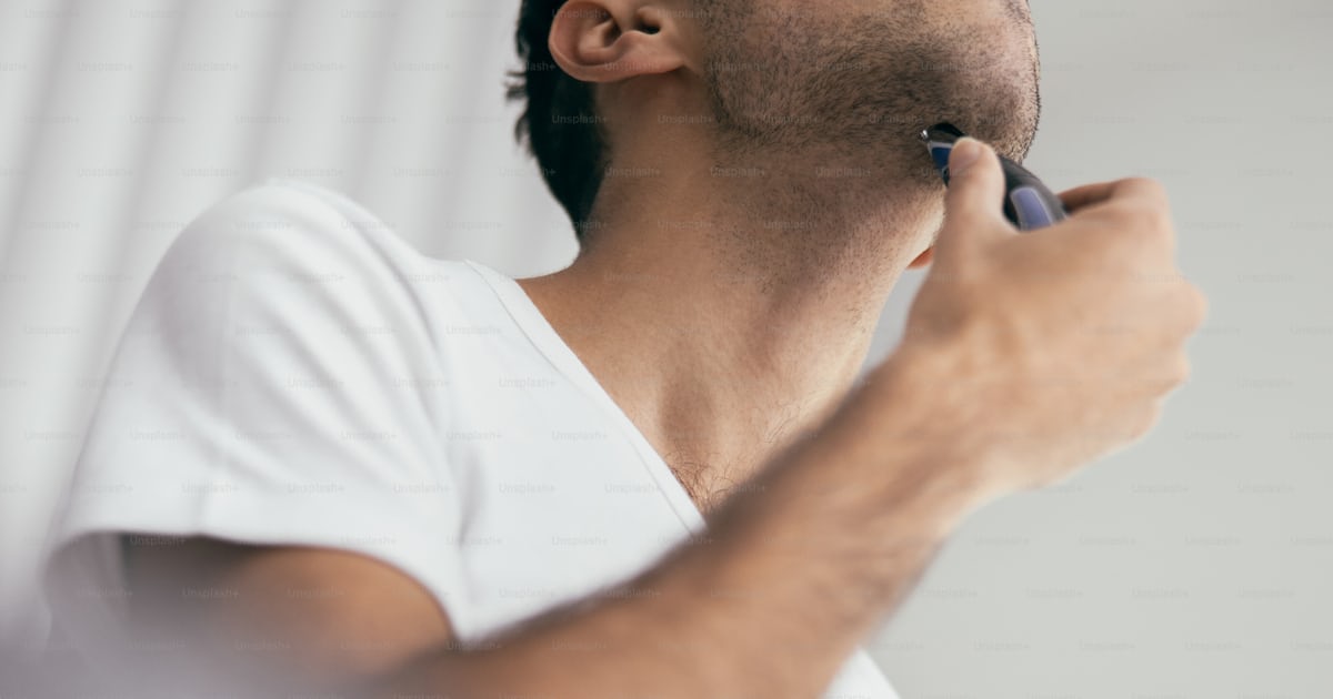 Close up of cheerful guy shaving beard with electric razor at home ...