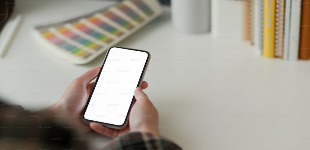Foto recortada de una diseñadora usando un teléfono inteligente de maqueta mientras trabaja con suministros de diseñador en una mesa blanca