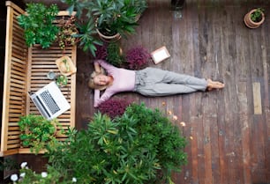 A top view of senior woman with laptop lying outdoors on terrace, resting.