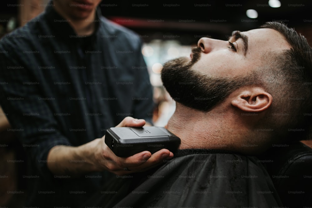 Hombre adulto joven guapo que recibe un tratamiento de peinado y vestimenta para el cabello y la barba por parte de un estilista profesional.