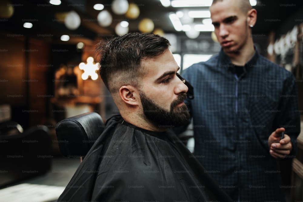 Gut aussehender junger Erwachsener, der von einem professionellen Friseur ein Haar- und Bartstyling und eine Verbandsbehandlung erhält.