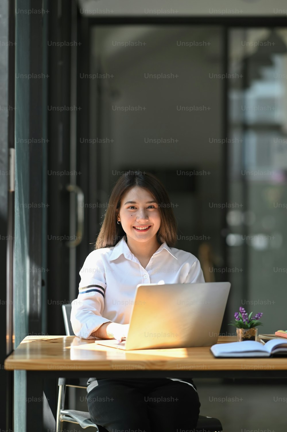 Photo of young beautiful woman smiling while sitting in front her computer laptop at the working wooden table over modern office as background.
