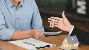 Immagine ritagliata del broker che dà una chiave al suo cliente che gli restituisce i soldi al tavolo da lavoro in legno con appunti e modello di casa che lo mette sopra.