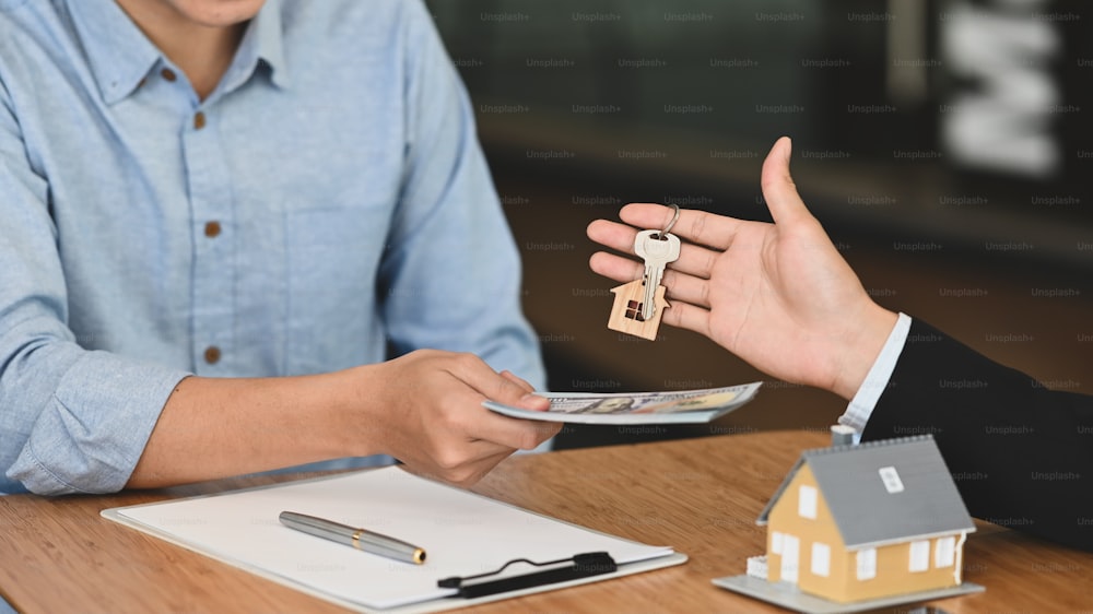 Immagine ritagliata del broker che dà una chiave al suo cliente che gli restituisce i soldi al tavolo da lavoro in legno con appunti e modello di casa che lo mette sopra.