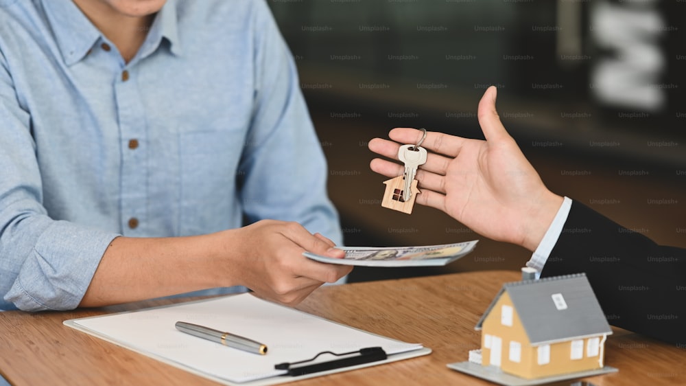Cropped image of broker giving a key to his customer that giving back money to him at the wooden working table with clipboard and house model putting on it.