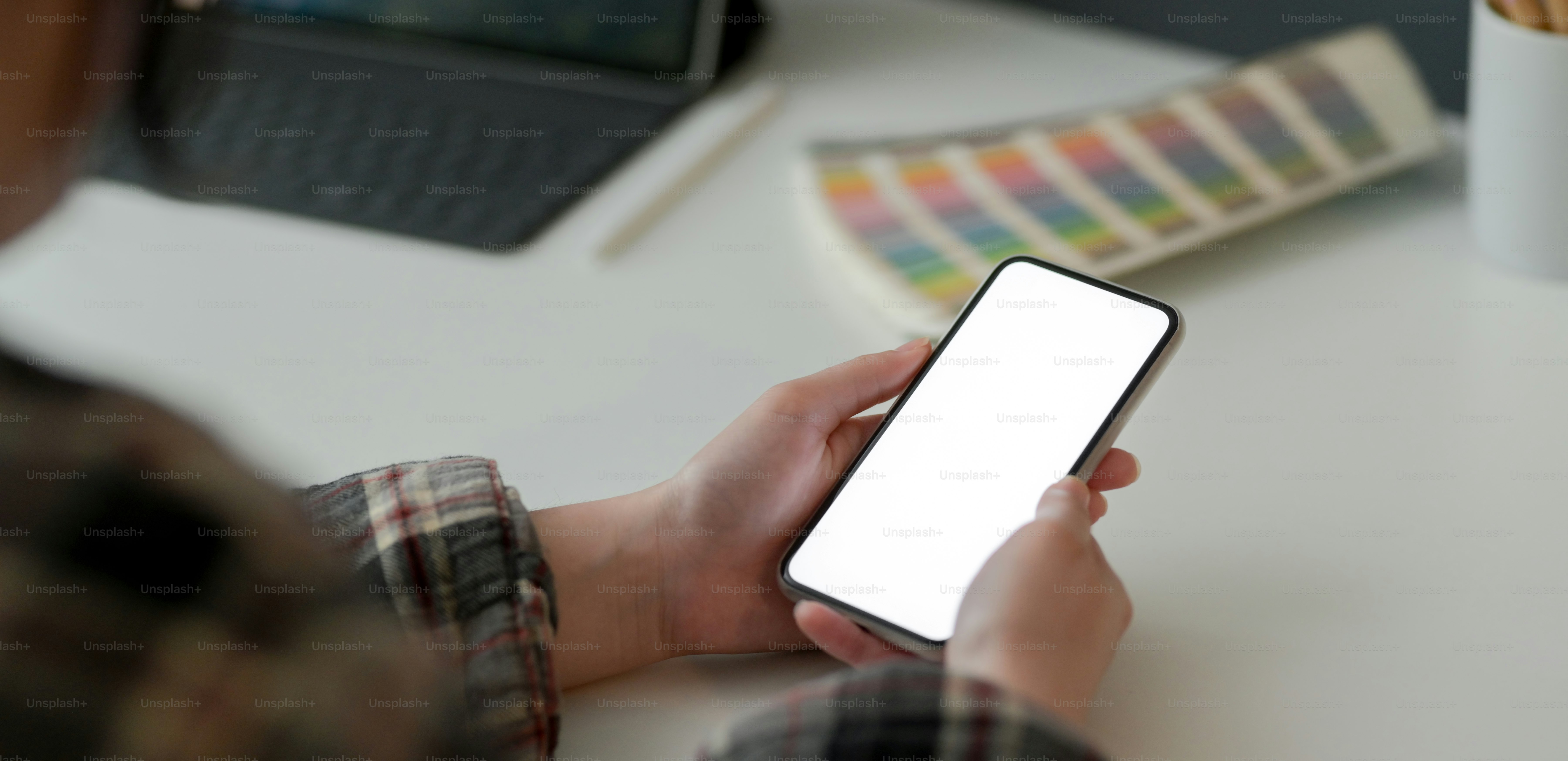 Colpo ritagliato di designer femminile che tiene in mano uno smartphone mock-up mentre lavora con tablet e forniture di design su tavolo bianco