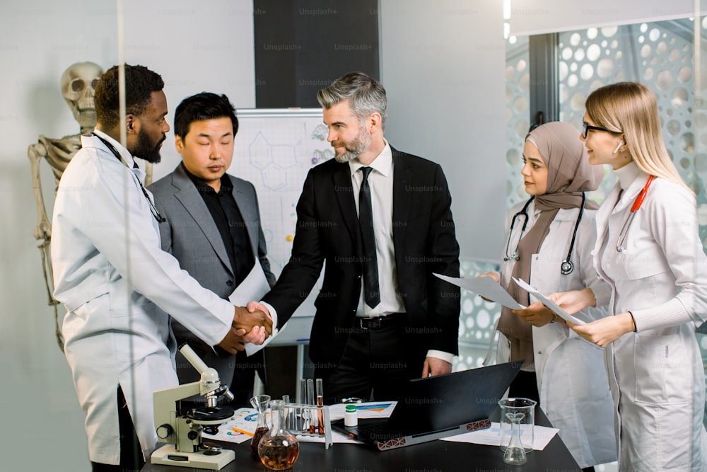 Mature Caucasian businessman and African man doctor shaking hands for some agreement, while standing together with their colleagues doctors, scientists, in modern lab office.