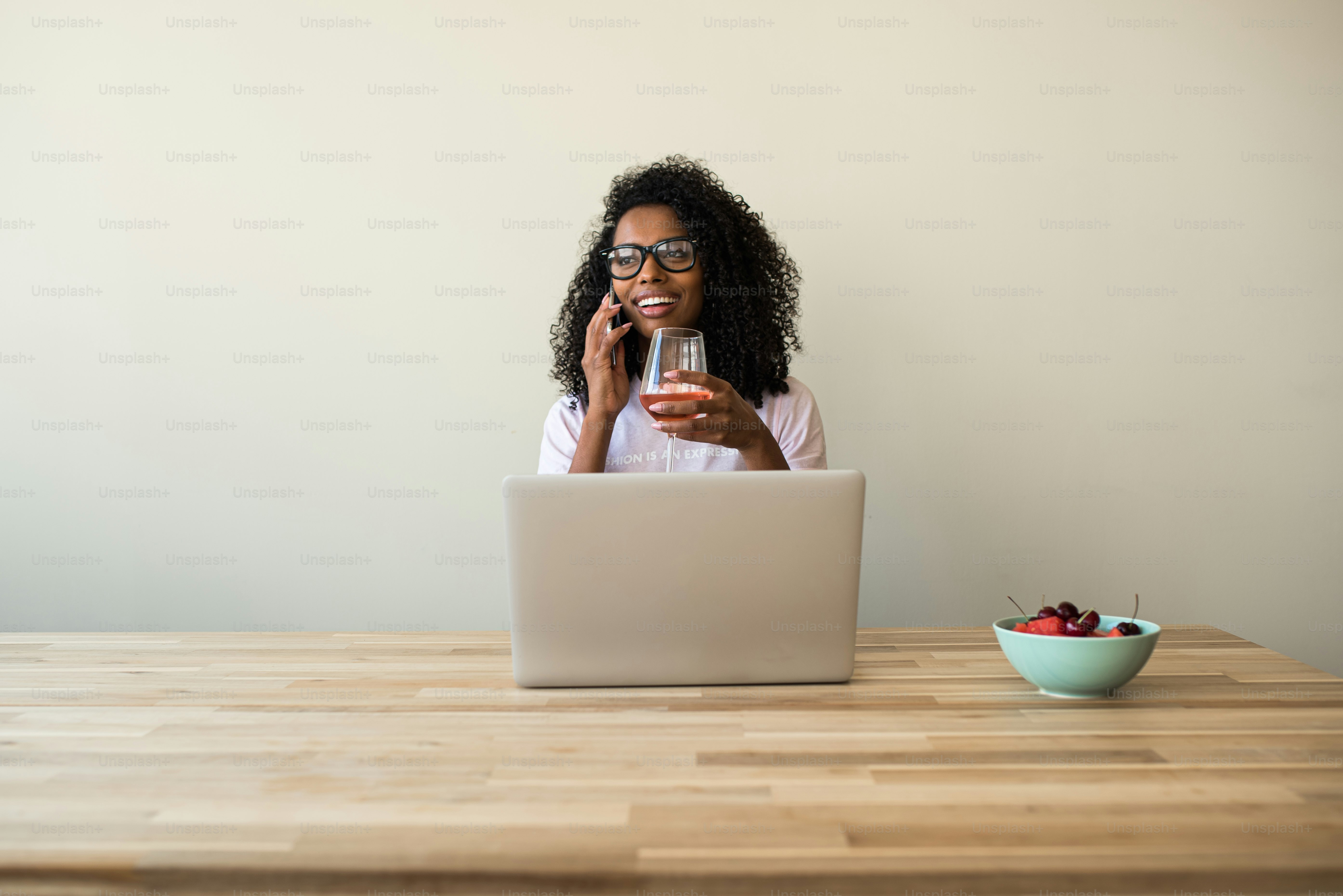 Focused black lady with curly hair browsing laptop ans using smartphone at wooden table with glass of wine and bowl with berries at home