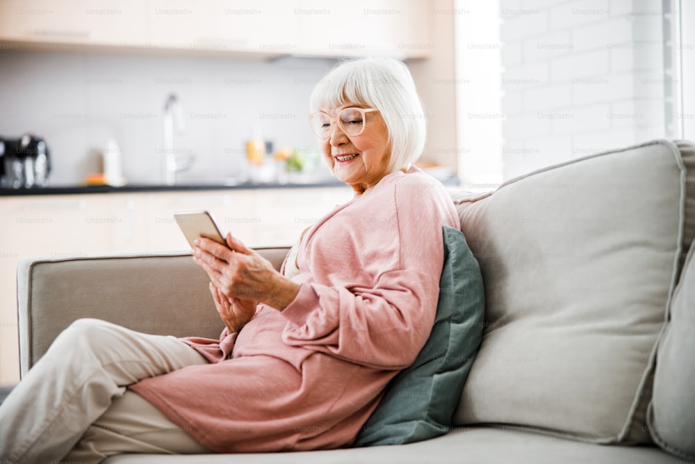 Lächelnde ältere Frau ruht sich auf dem Sofa aus und liest Nachricht auf Smartphone-Stockfoto
